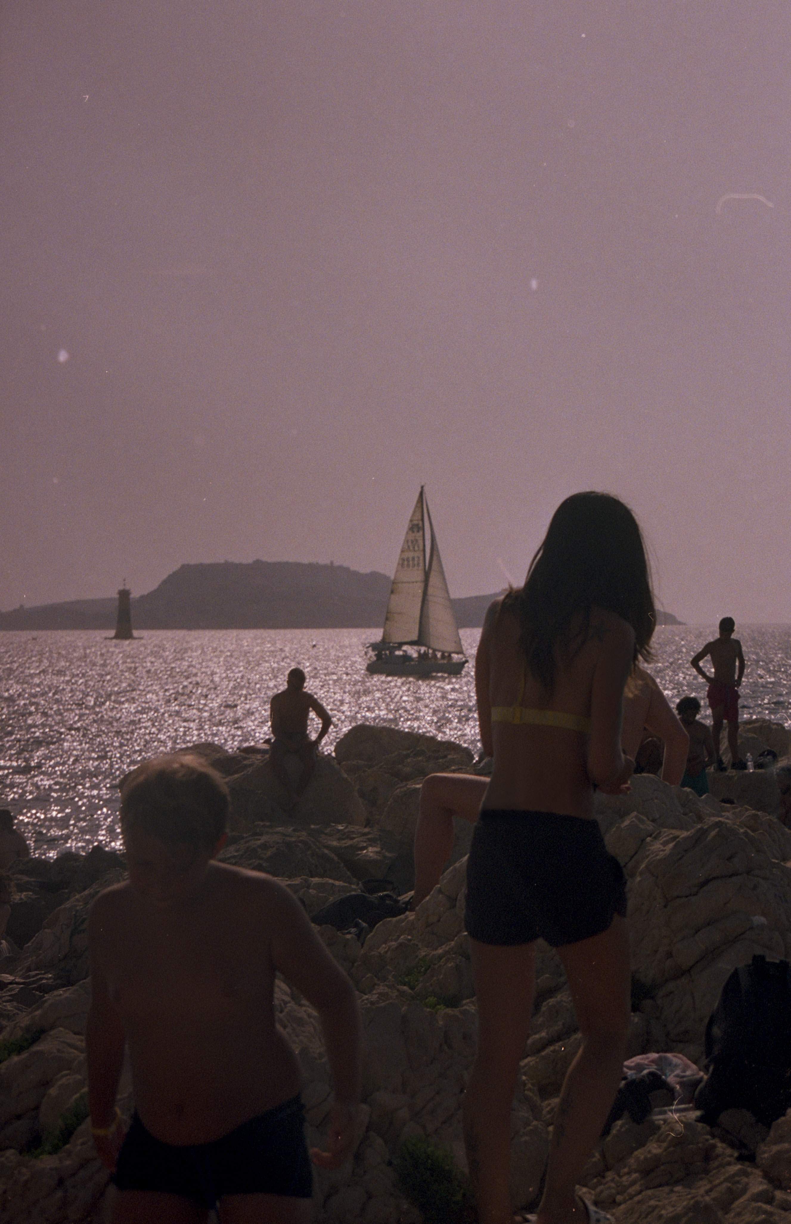 People sat and walking on rocks by the sea, sailing boat in the background