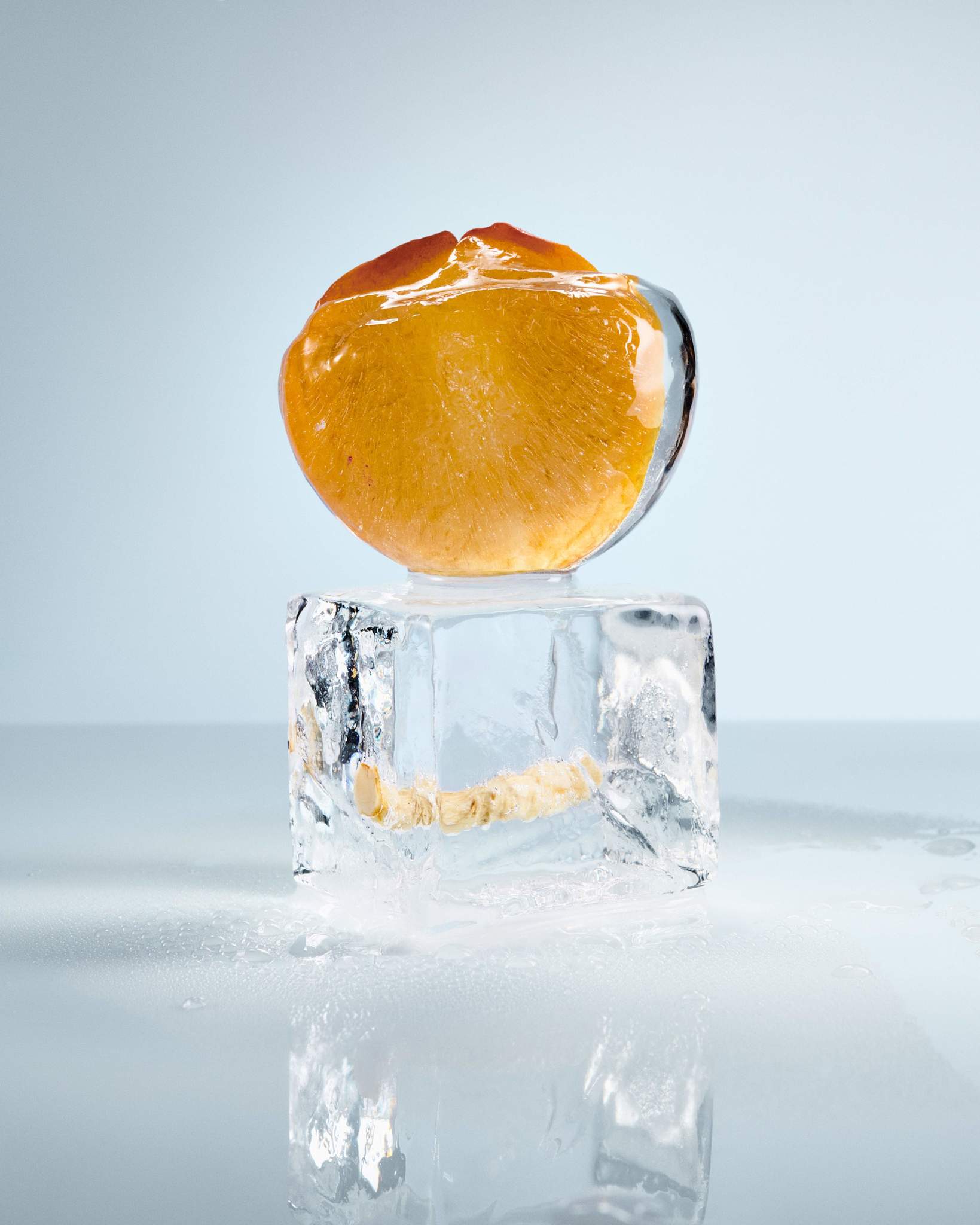 Sliced apricot perched on a clear, melting ice cube, with reflections and water droplets on a light blue backdrop.