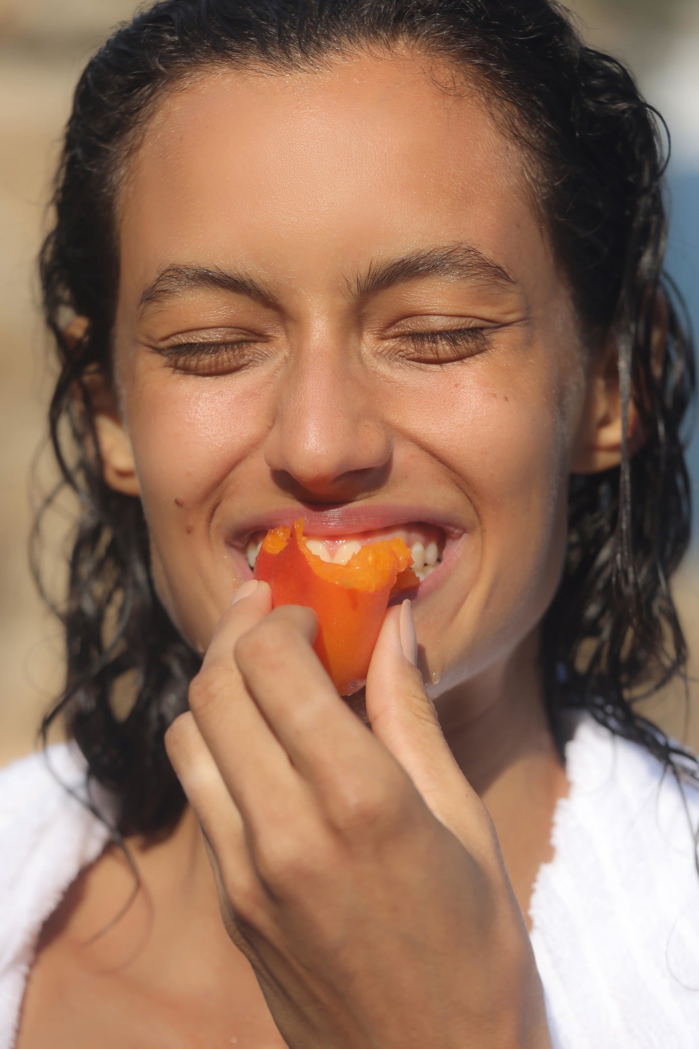 Woman at the beach, smiling with her eyes closed and biting into a fresh apricot. Her hair is wet and the sun is shining.